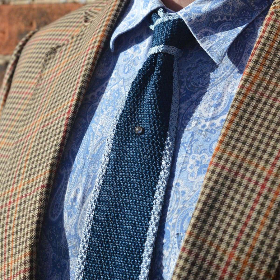 Close-up of a man's outfit featuring a blue knit tie, plaid blazer, and paisley shirt showcasing classic formal style.