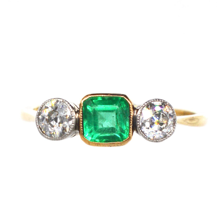 Elegant Edwardian three stone ring featuring a vibrant emerald and sparkling diamonds in gold and platinum setting.