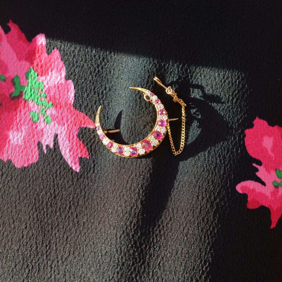 Edwardian 18ct gold crescent moon brooch with ruby and diamond accents against floral background.