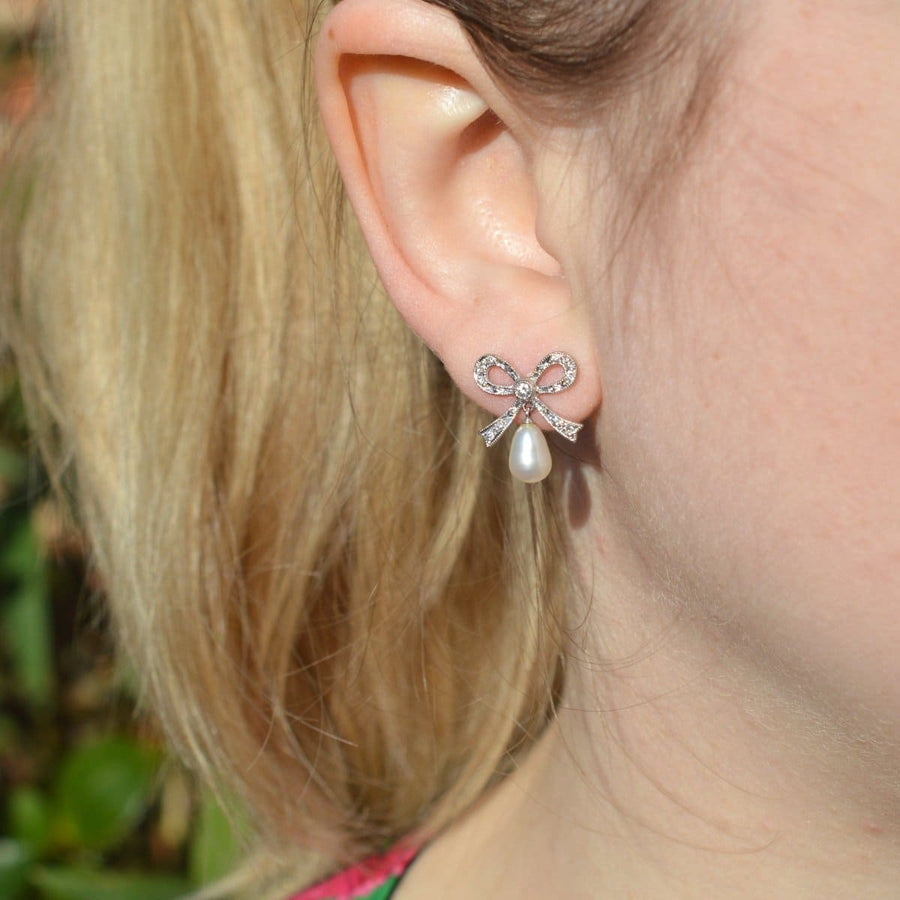 Vintage 18ct white gold diamond bow and cultured pearl drop earrings worn on a model's ear outdoors.